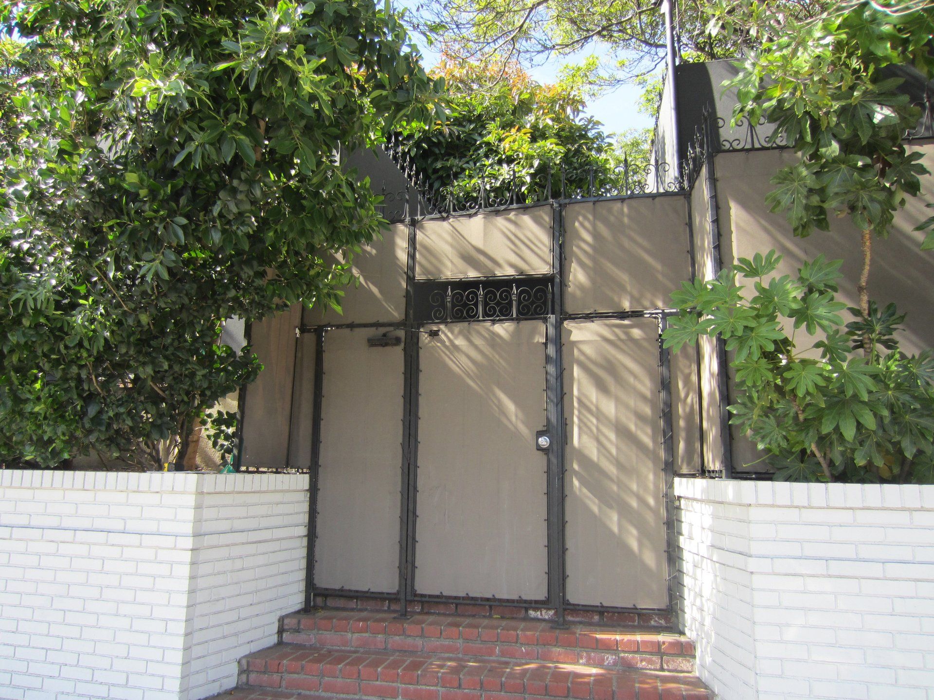 Front gate with beige panels and black trim, framed by white brick walls and leafy trees.