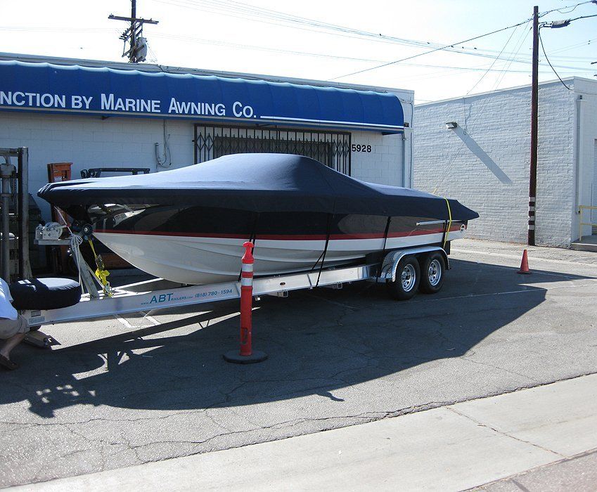 Covered boat on trailer parked outside a marine awning shop, with orange cones nearby.