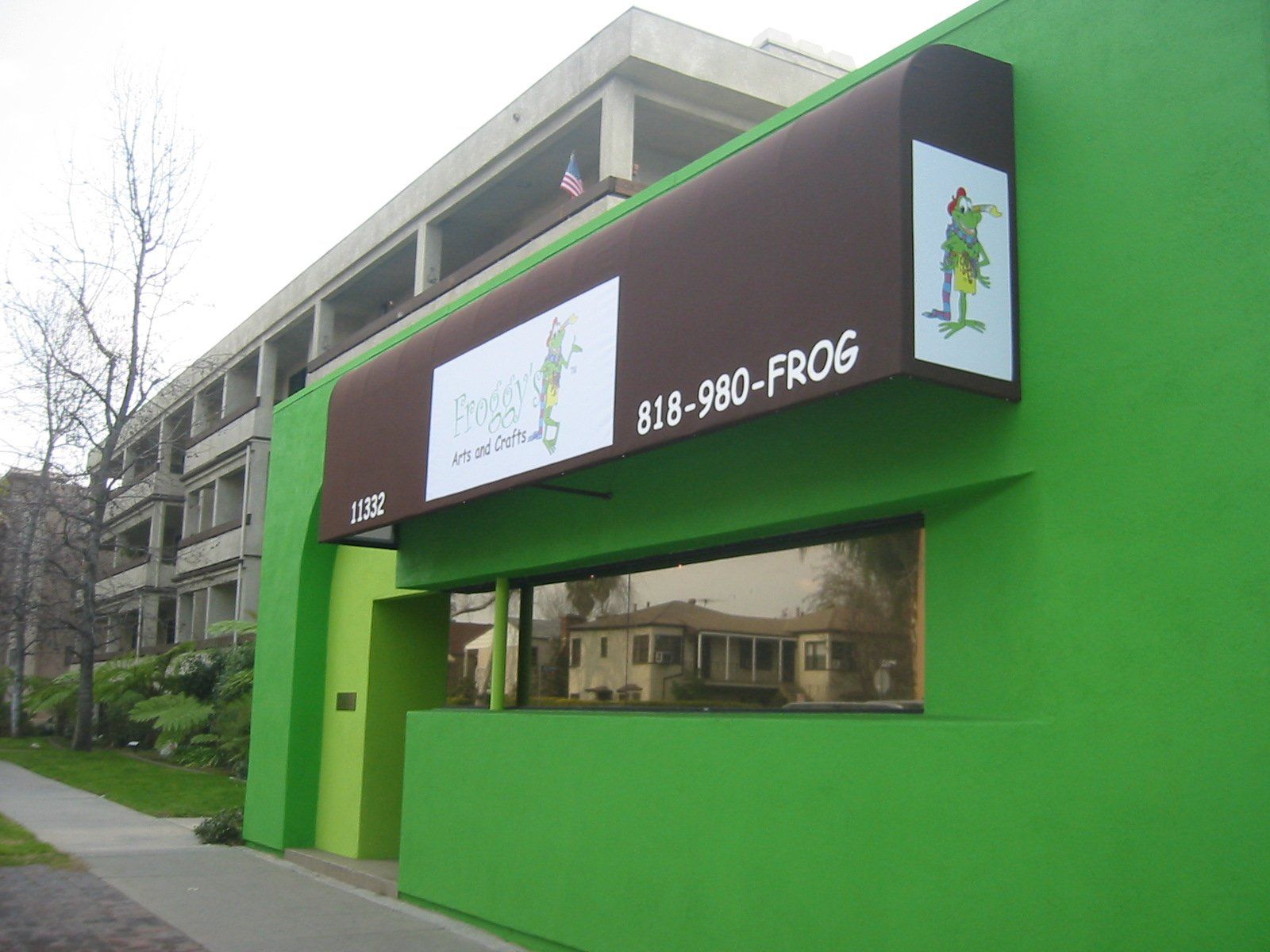 Green storefront with a black awning labeled “FROG” beside a sidewalk and apartment building.