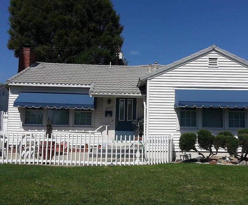 White house with blue awnings, a white picket fence, and a green lawn under a clear blue sky.