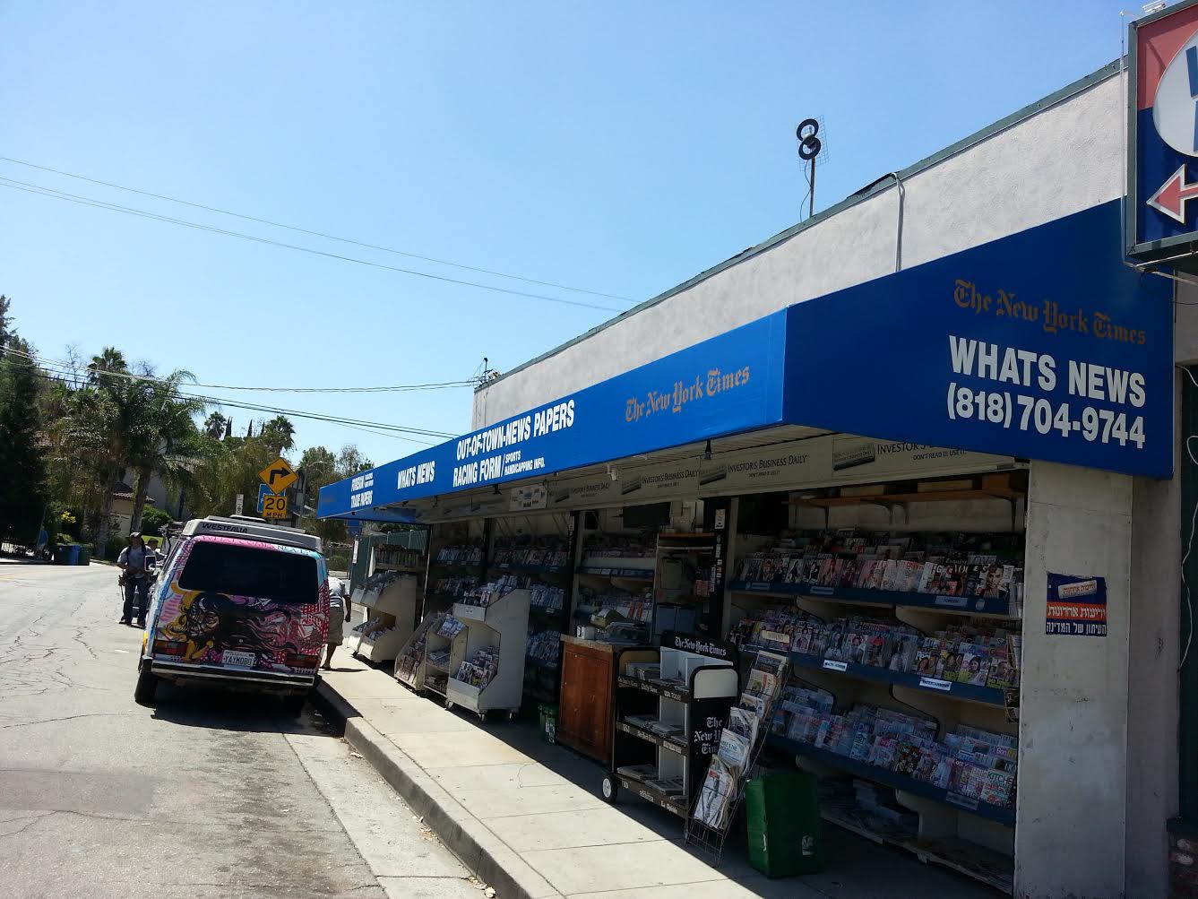 Small roadside newsstand with blue awning, magazines outside, and a van parked along the curb