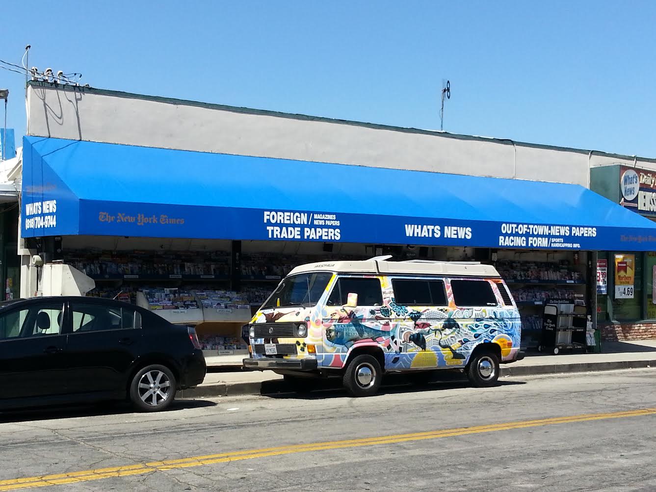 Storefront with blue awning and a decorated white van parked in front, beside a black car on the street