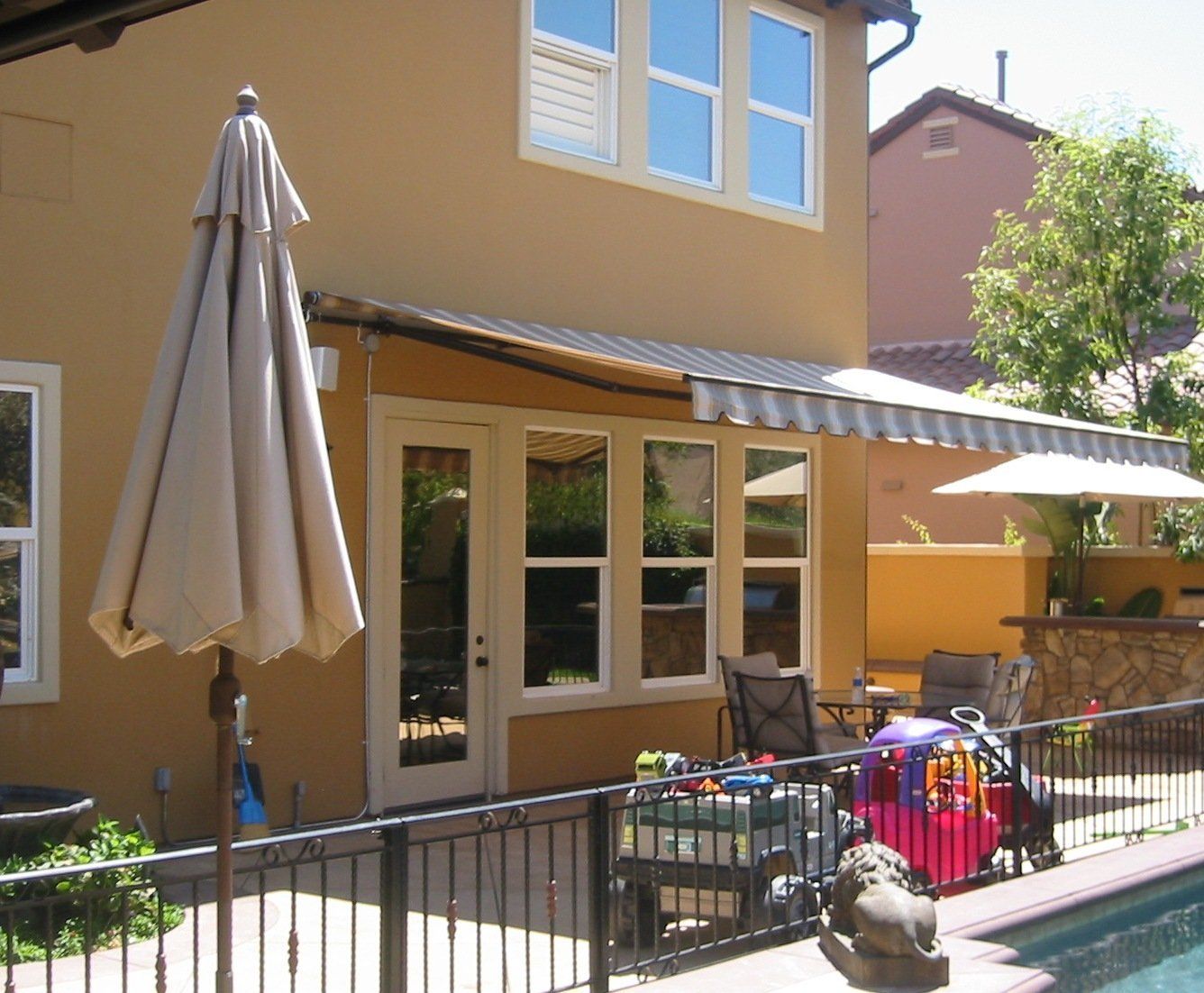 Backyard patio with table, umbrella, chairs, pool fence, and house exterior in sunlight