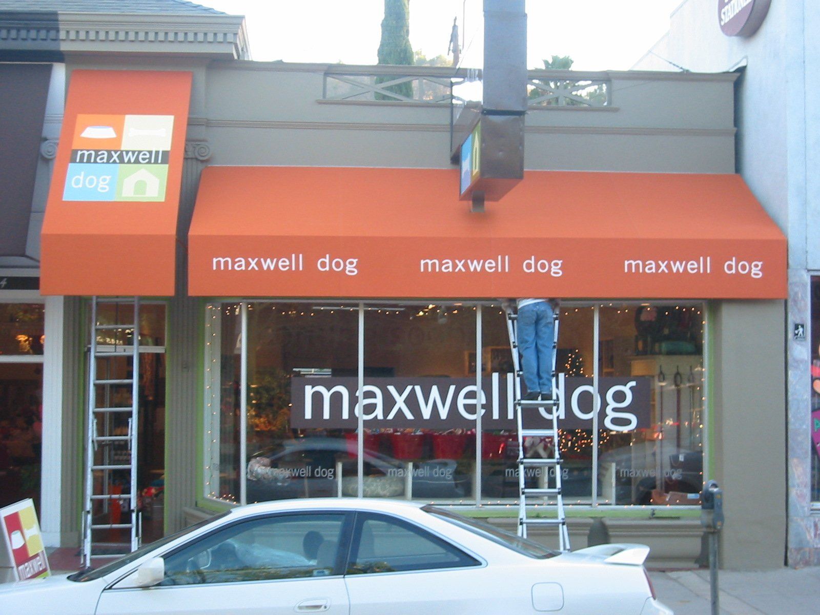 Storefront with orange awning and “maxwell dog” signage above a parked silver car.