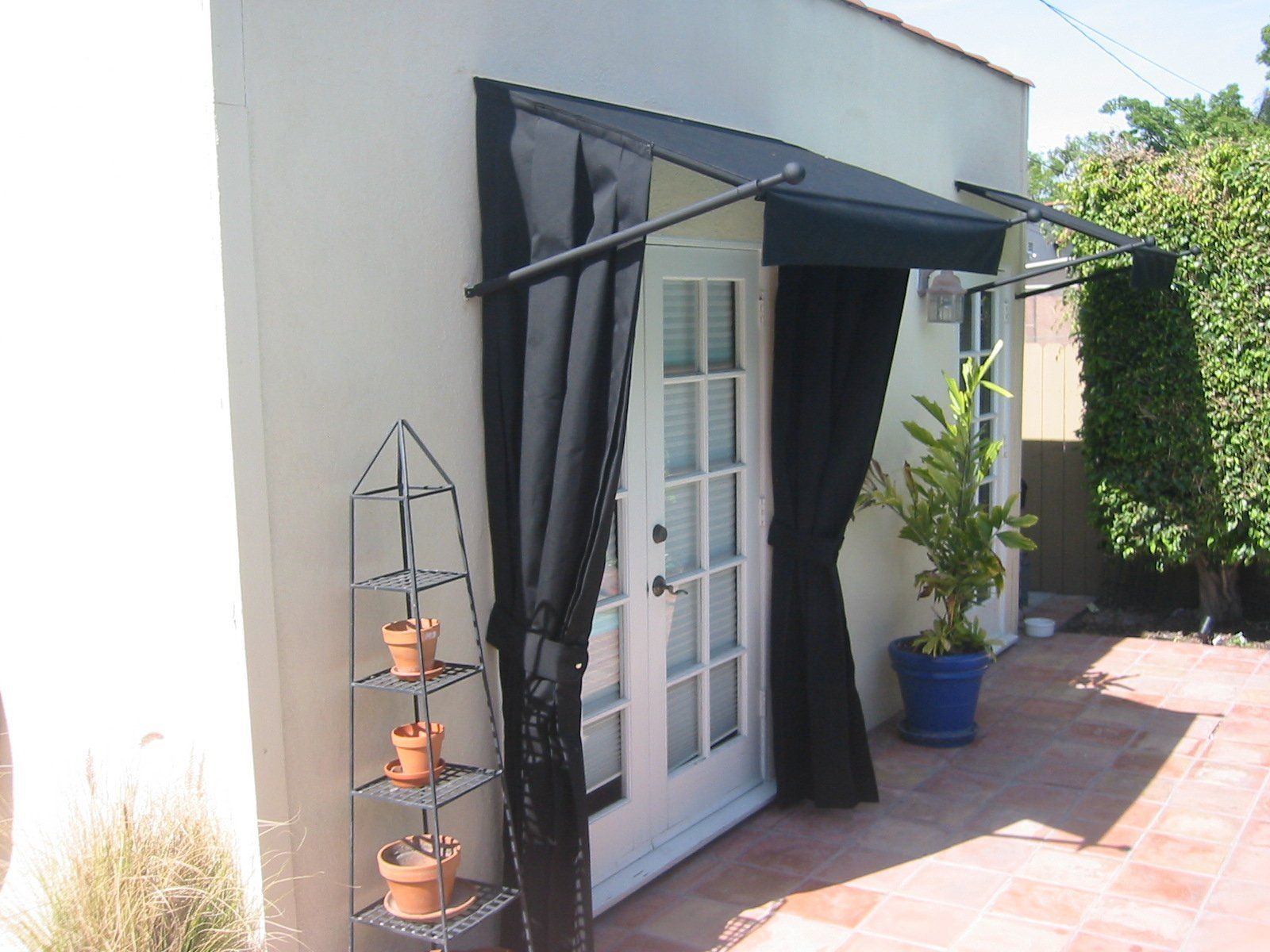 Patio door with black curtains, terracotta tile floor, and potted plants beside a white wall