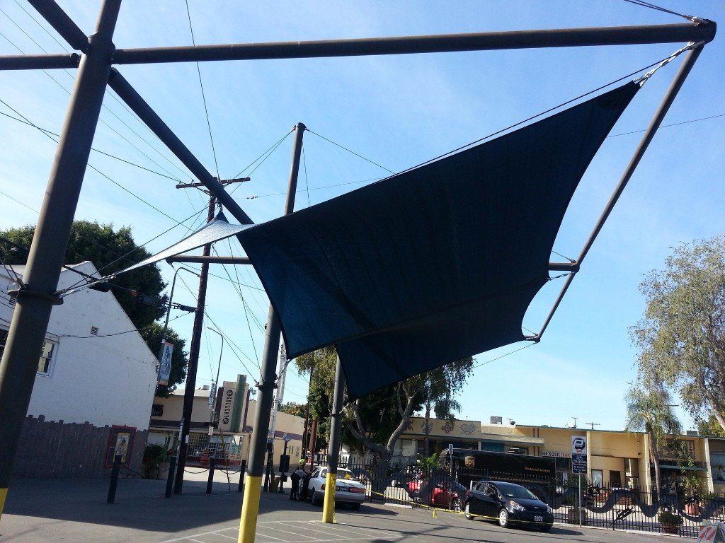 Large dark shade sail over a sunny outdoor plaza with trees, buildings, and parked cars.