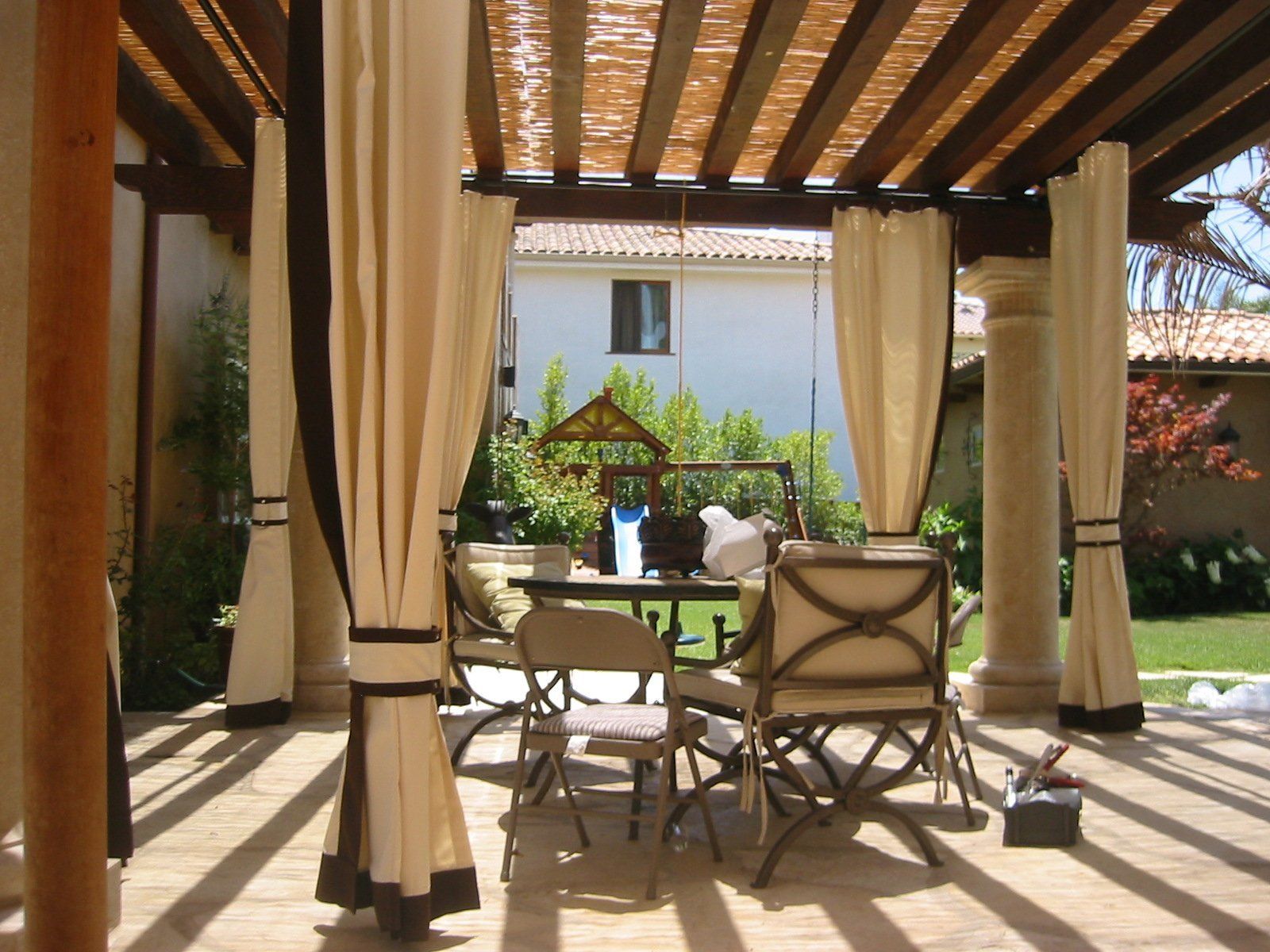 Covered patio with cream curtains, wicker chairs, and a small table in sunlight facing a garden courtyard.