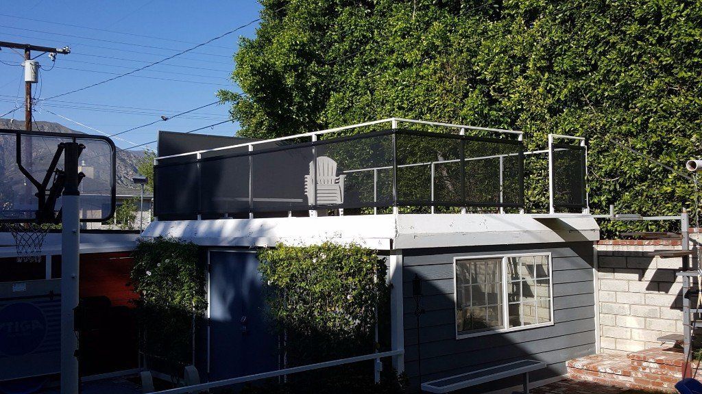 Backyard rooftop deck with black mesh railing on a small white outbuilding, beside a pool and trees.
