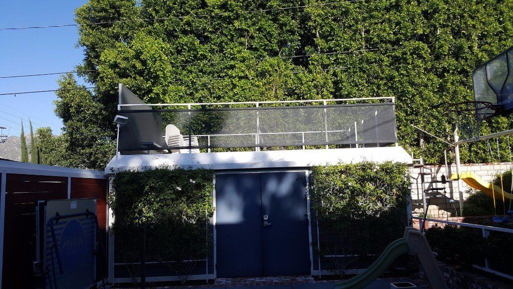 Small building with a blue door, rooftop railing, and dense green trees behind it under a clear sky