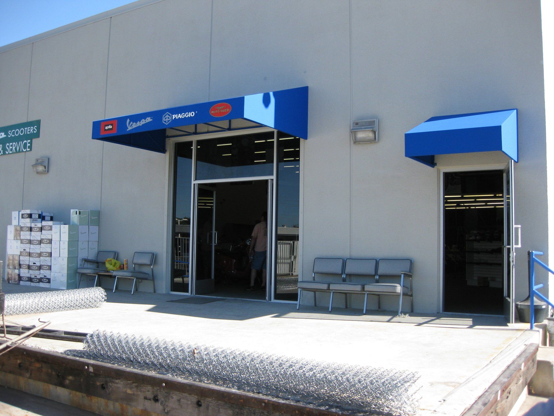Exterior of a blue storefront with open doors, benches, and a sunlit loading ramp