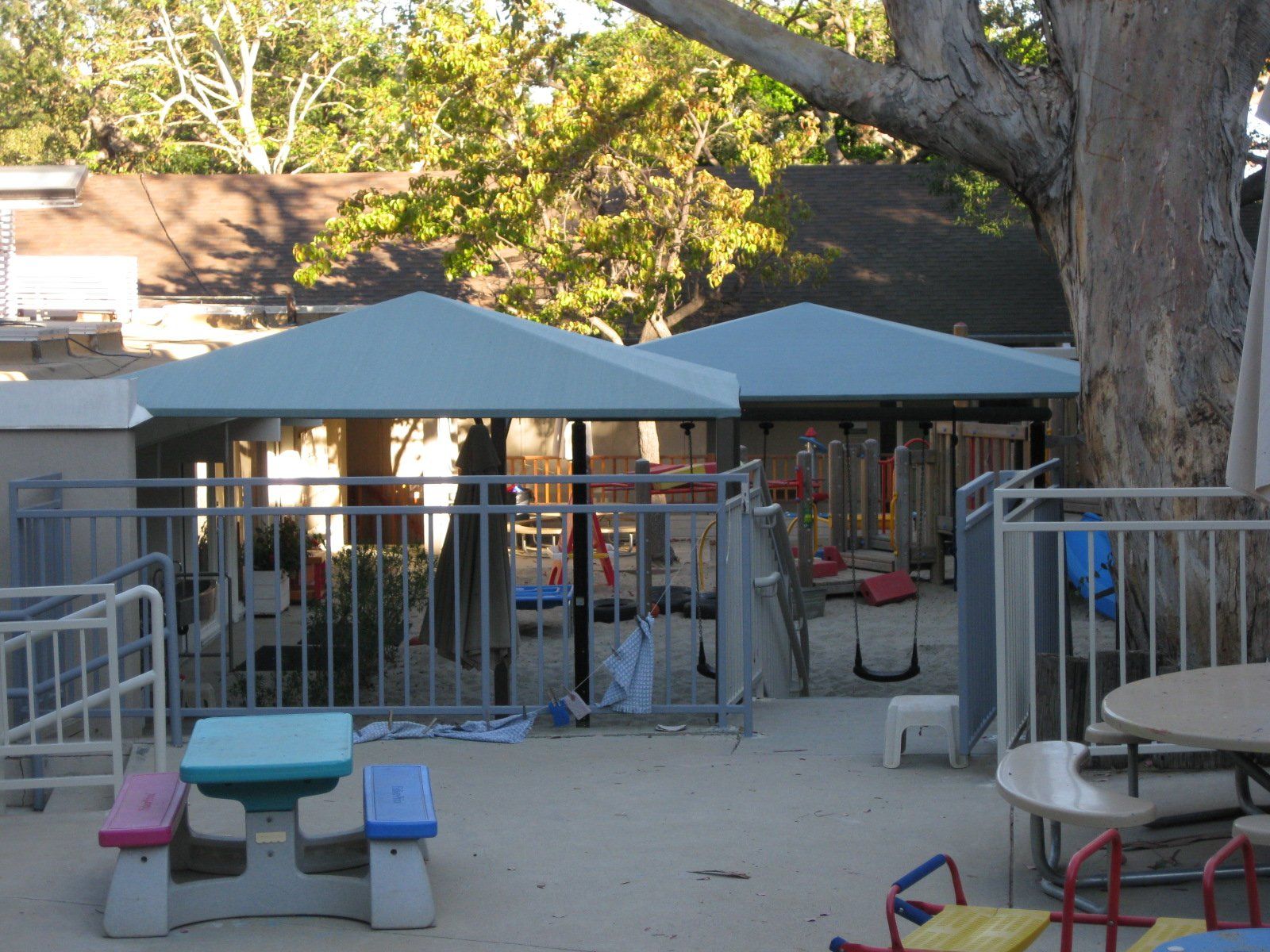 Outdoor playground with blue canopies, picnic tables, and metal railings under a large tree