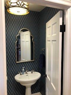 Small powder room with blue patterned wallpaper, pedestal sink, gold-framed mirror, and white door.