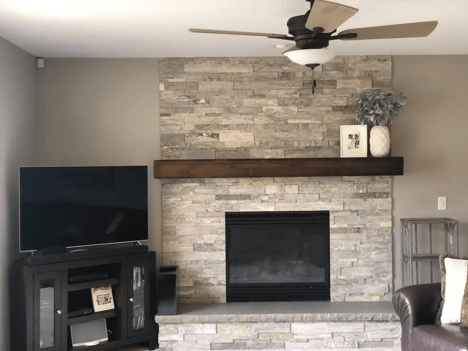 Living room with stone fireplace, TV, and ceiling fan. Gray walls, brown mantel, black TV stand.