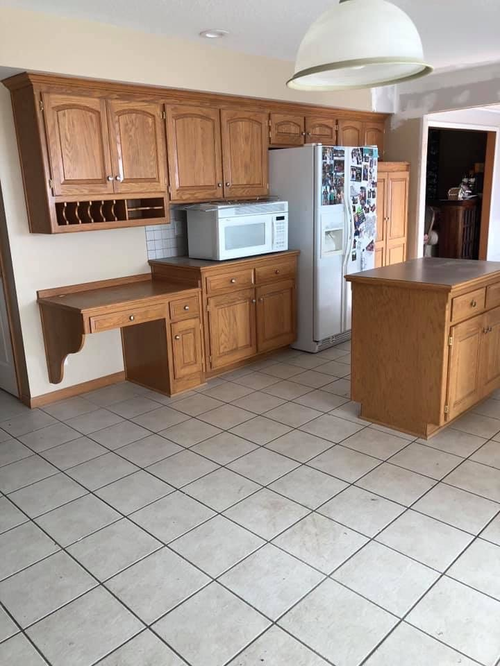 A kitchen with light wood cabinets, a microwave, refrigerator, island, and desk. Beige tile floor.