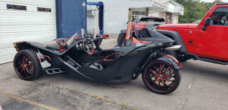 A black Polaris Slingshot three-wheeled vehicle with custom red and black wheels parked in an outdoor lot.