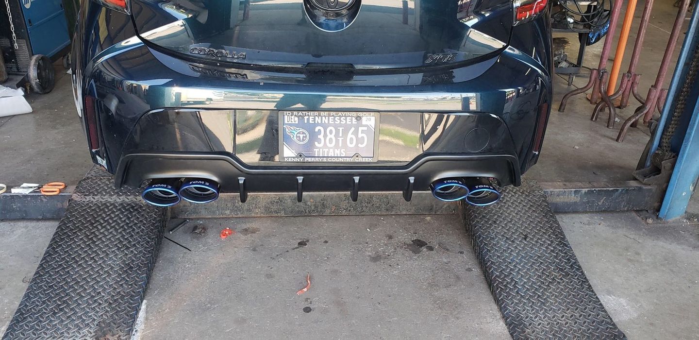 Rear view of a dark-colored sports car parked on vehicle lift ramps in a garage, featuring a quad-exhaust and diffuser.