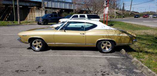 A gold 1968 Chevy Chevelle with a white vinyl roof parked in an outdoor lot near a bridge.