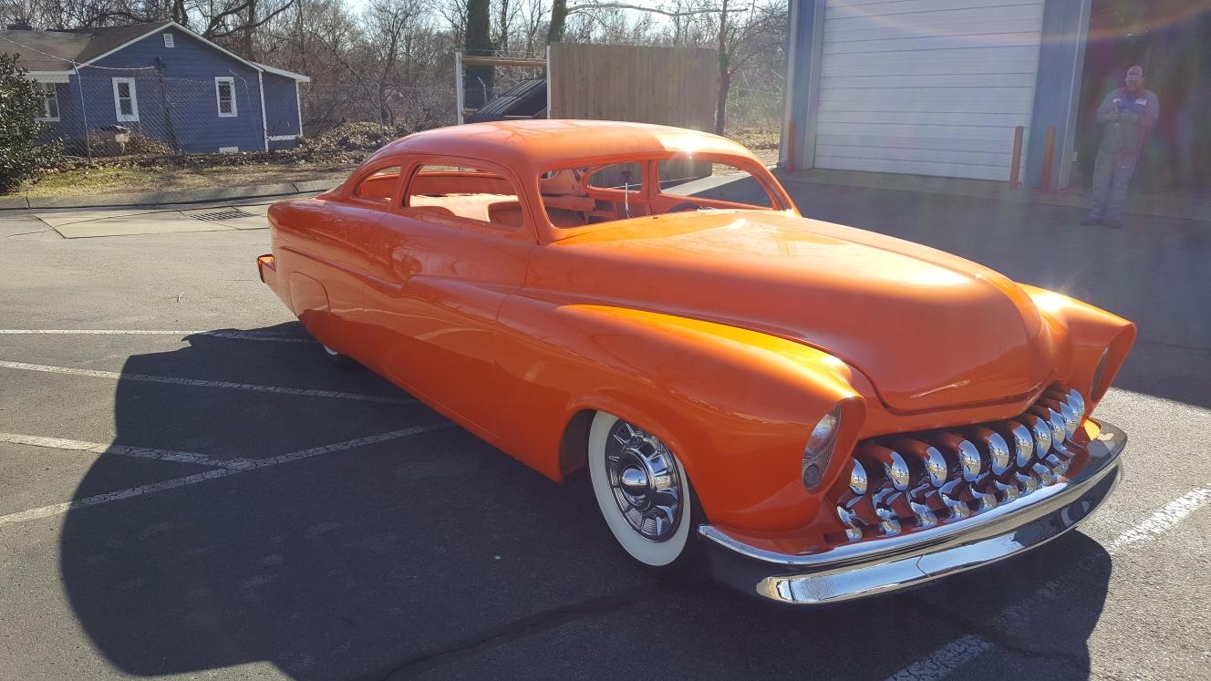 A bright orange, customized vintage car with a prominent chrome grille and whitewall tires parked on an asphalt lot.