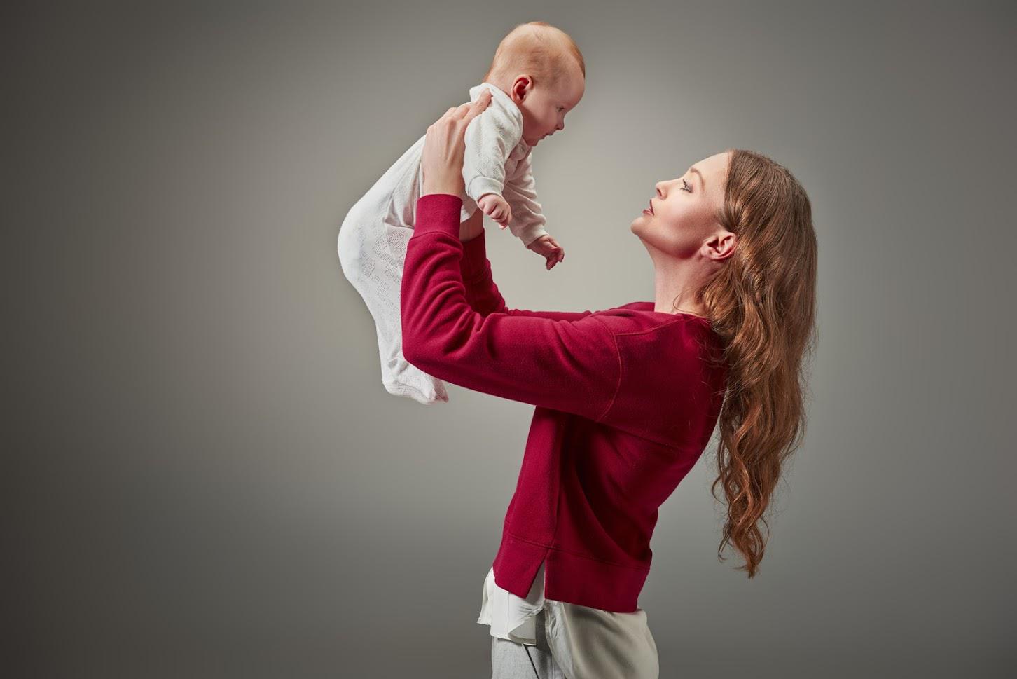 Woman in red sweater holds baby in white onesie up, looking at it. Neutral gray background.