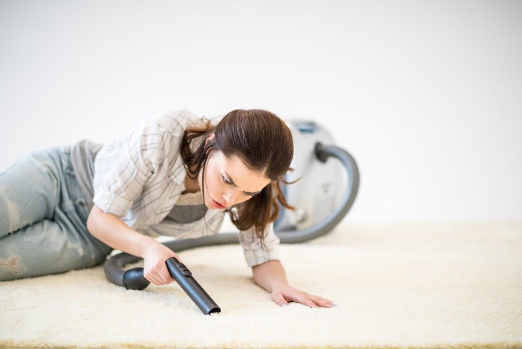 Woman on hands and knees vacuuming a white carpet. Vacuum cleaner in background, white wall.