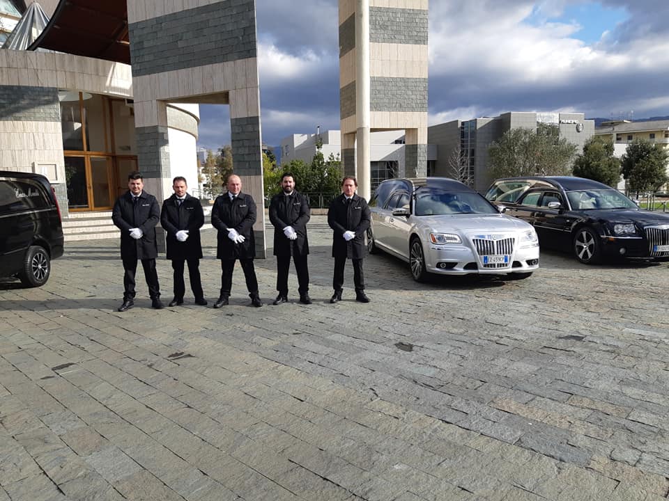 Cinque persone in piedi accanto ad auto parcheggiate in una piazza pavimentata con alte colonne moderne e un cielo nuvoloso.