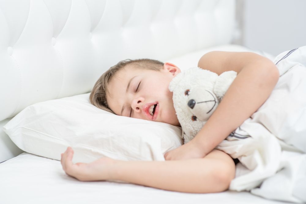 Child sleeping on a white pillow under white sheets while clutching a white teddy bear.