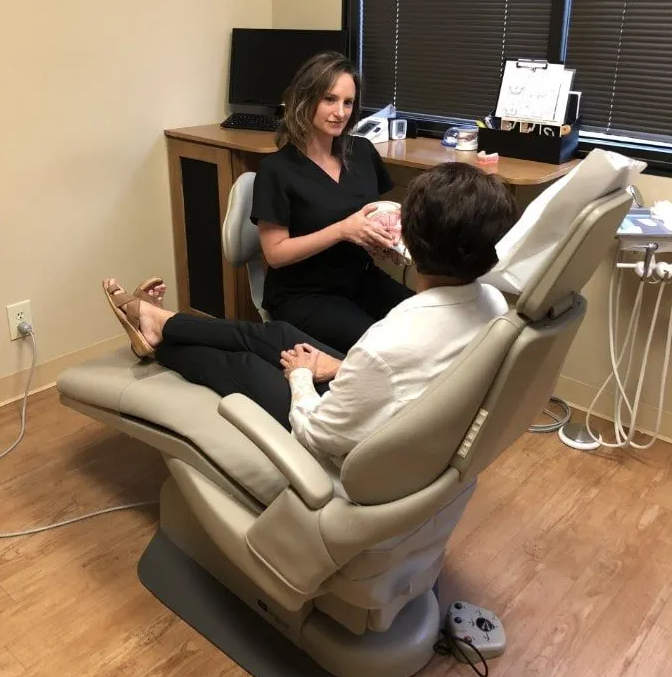 Female doctor in black scrubs speaks with female patient in white blouse sitting in patient's chair.