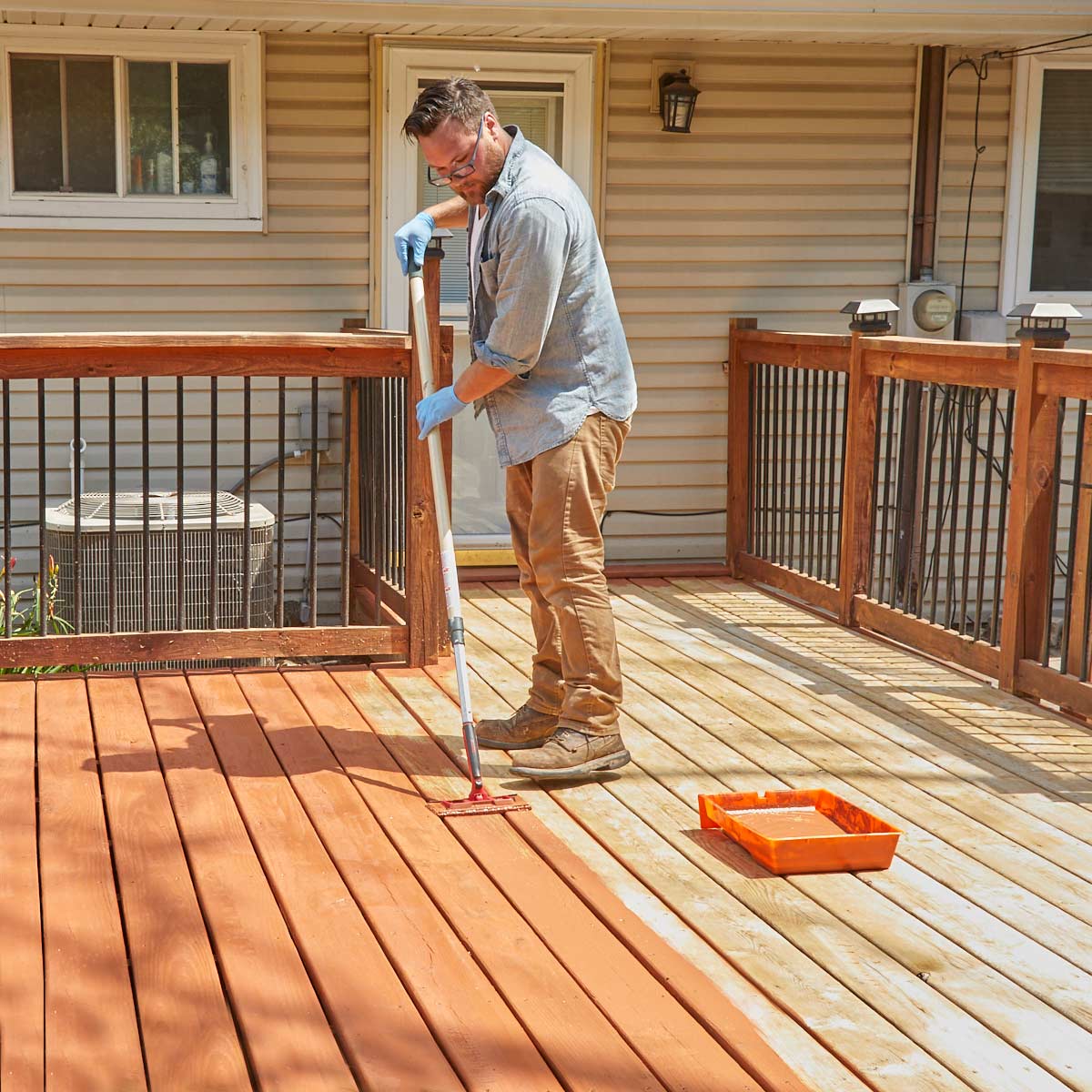 Man staining a wooden deck with a roller, wearing gloves and work boots. Sunny outdoor setting.