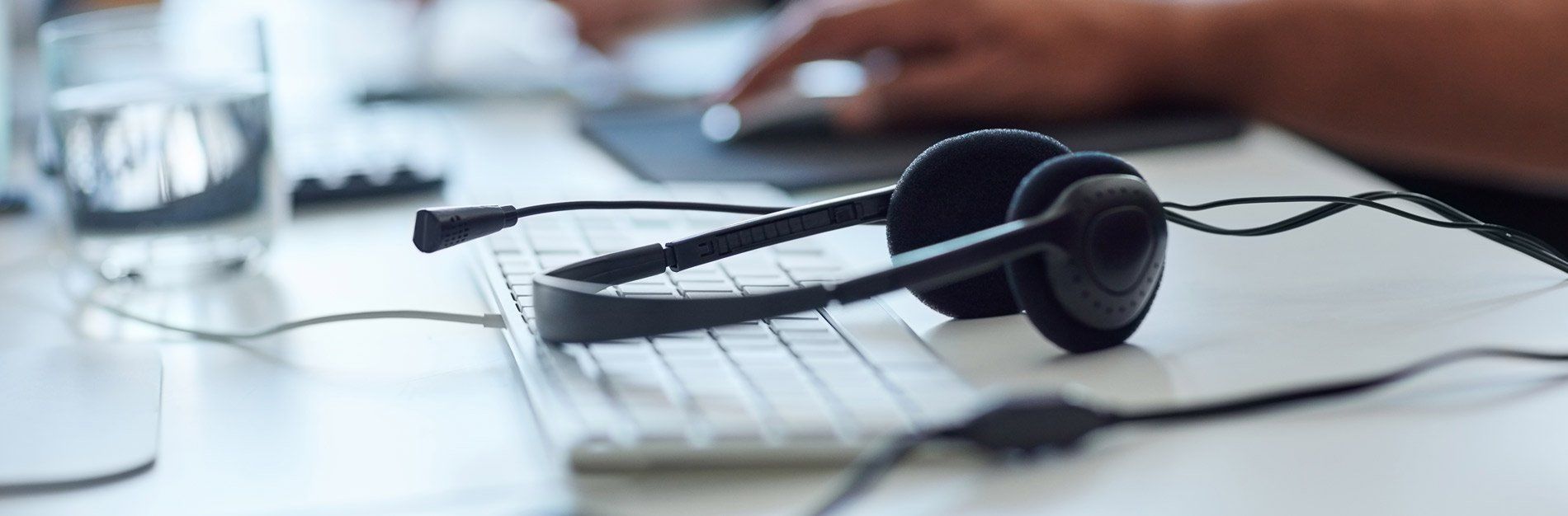Headset lying on a keyboard, person using a computer in the background.