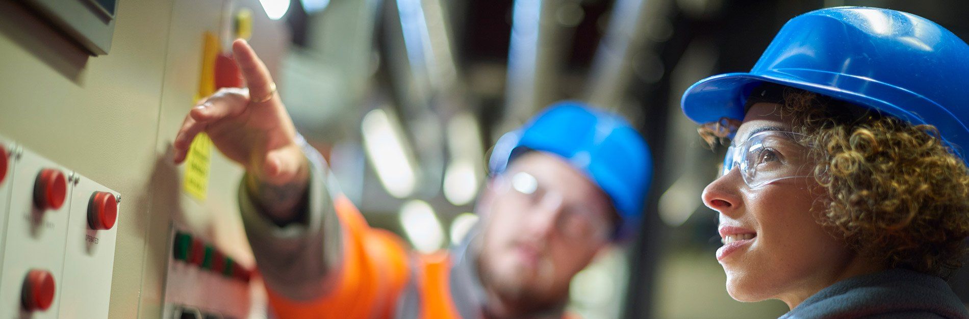 Two workers in blue hard hats and safety glasses inspect equipment. One points at a panel.