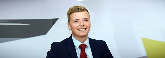 Man in a suit smiling at the camera, with a background of white and yellow geometric shapes.