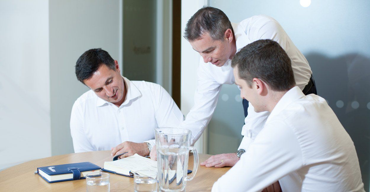 Three men in white shirts, around a table, looking at a notepad. Office setting, focused.