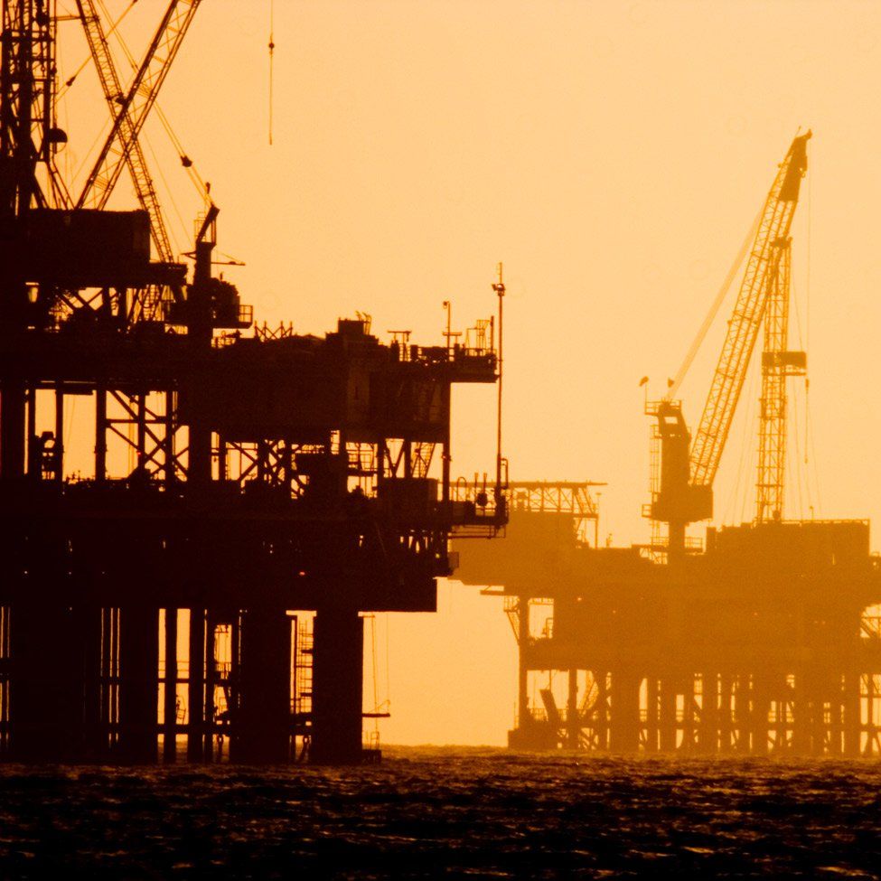 Silhouette of two offshore oil rigs against a golden sunset over the ocean.