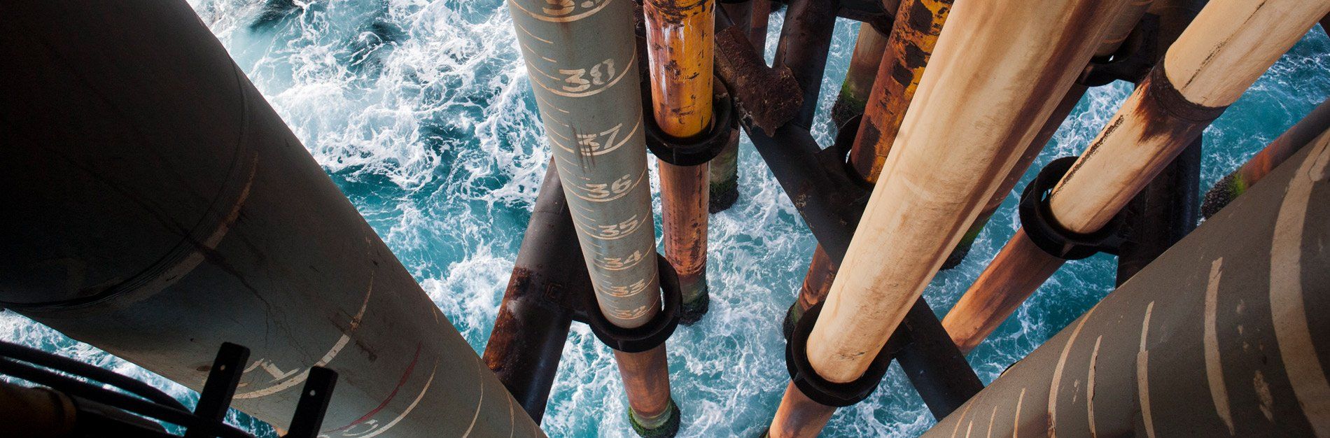 Overhead view of an offshore oil rig with various pipes extending down into the blue ocean water.