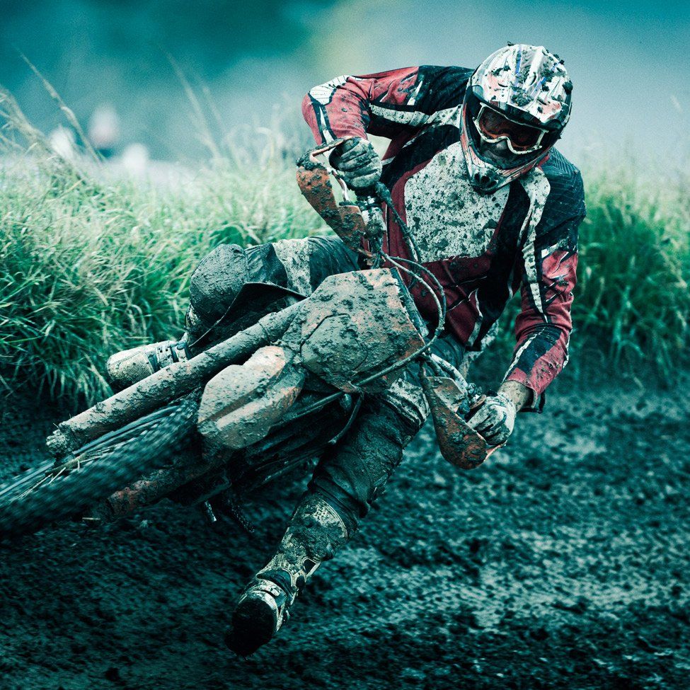 Motorcyclist on a muddy dirt bike track, covered in mud, leaning into a turn.