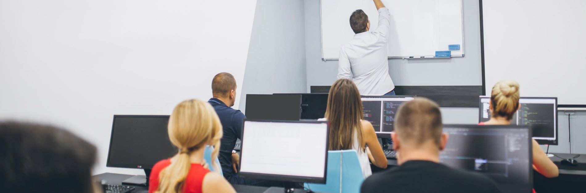 Students in a classroom listen to a lecture. A man writes on a whiteboard while students work at computers.