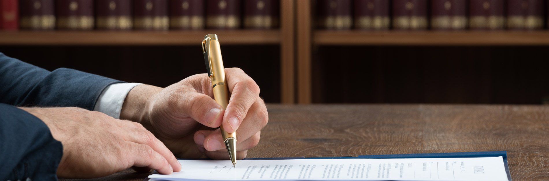 A person's hand with a gold pen writing on paper, possibly signing a document, with bookshelves in the background.