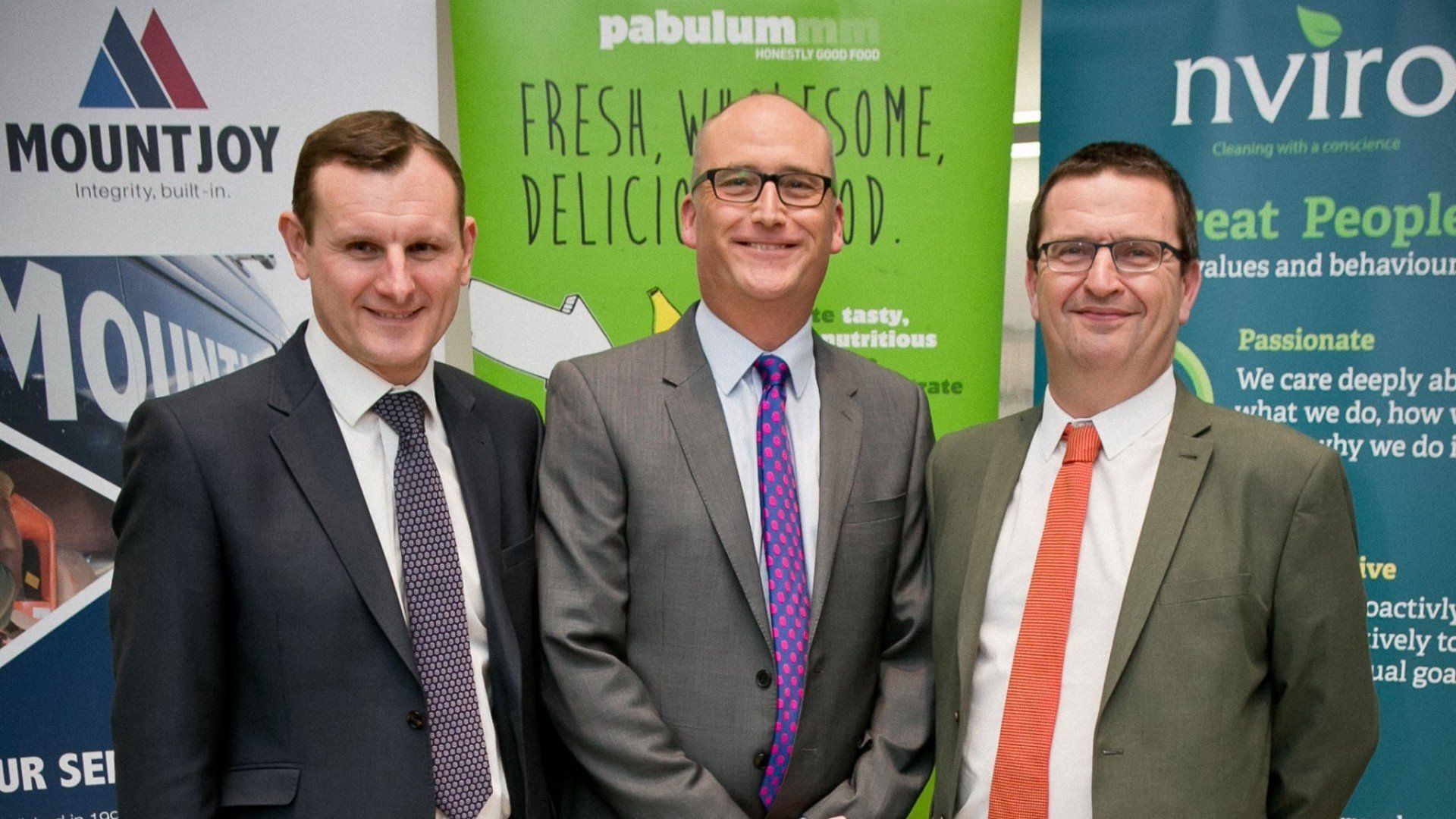 Three men in suits stand in front of company banners.