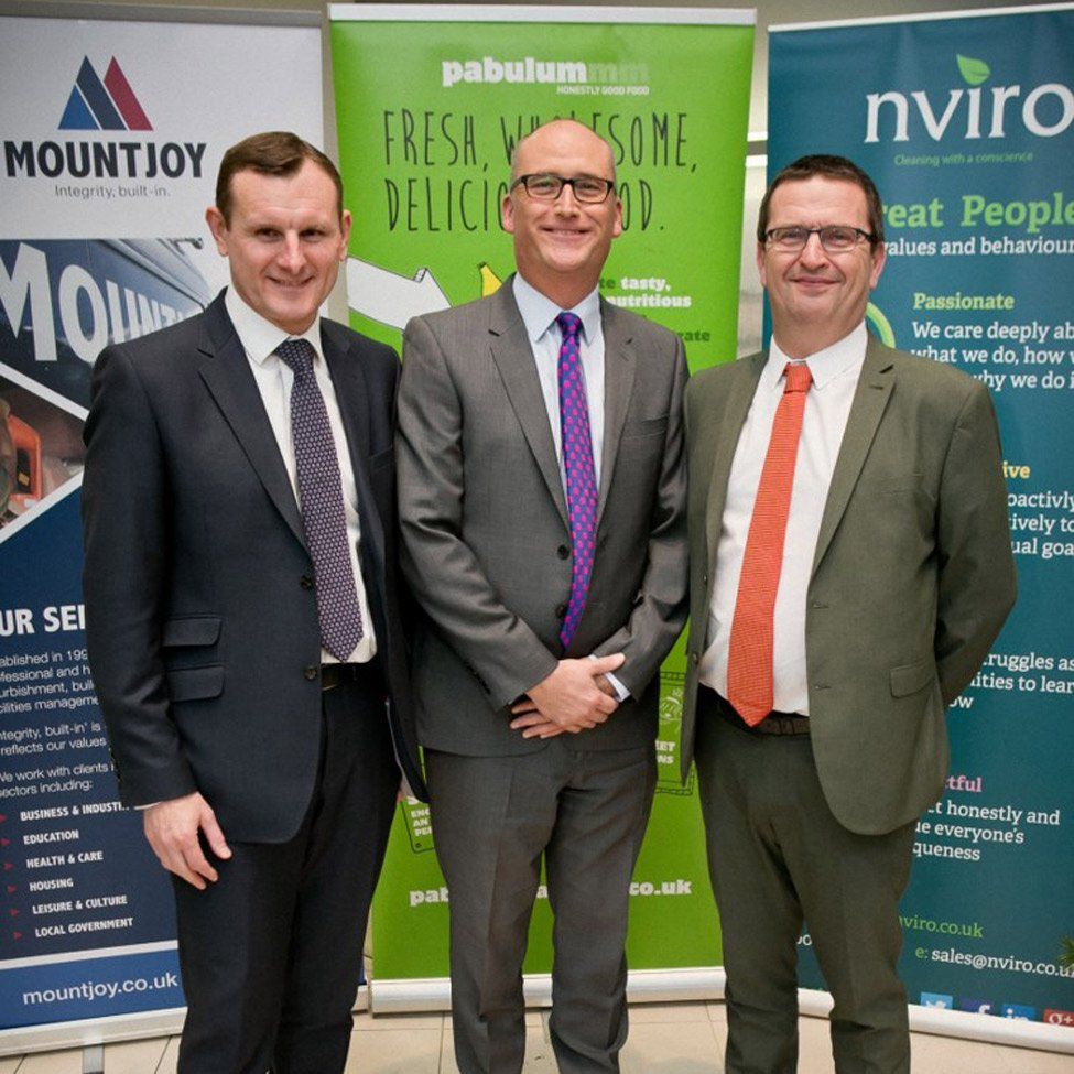 Three men in suits stand in front of banners. One has a purple tie, another an orange tie.