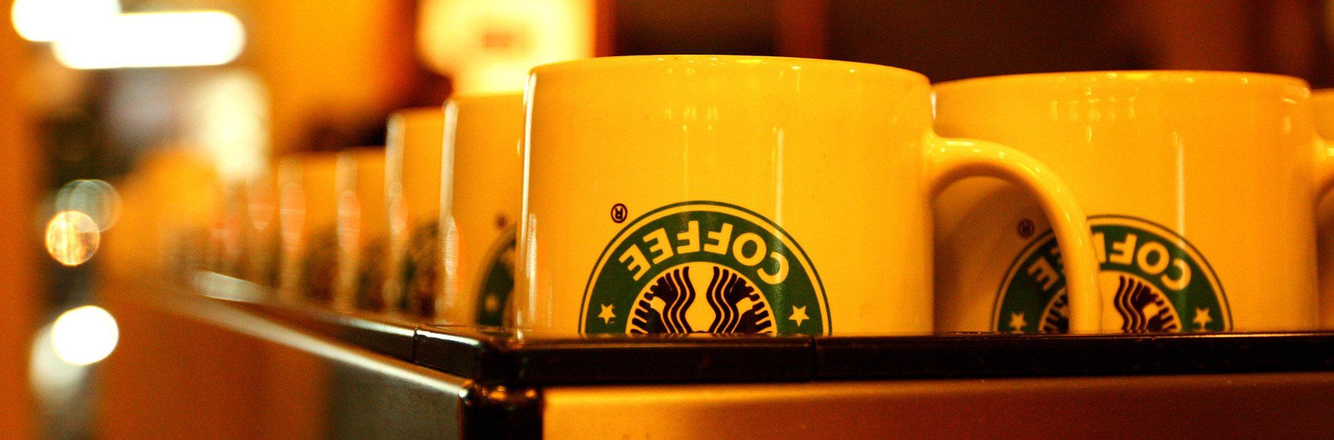 Row of Starbucks coffee mugs. Mugs are yellow with a Starbucks logo, lined up on a dark shelf.