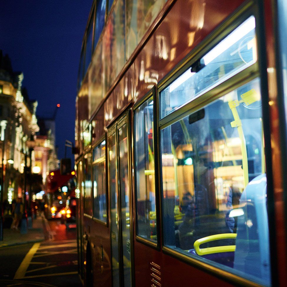 Red double-decker bus on a city street at night. Lights reflect in the windows; buildings in the background.