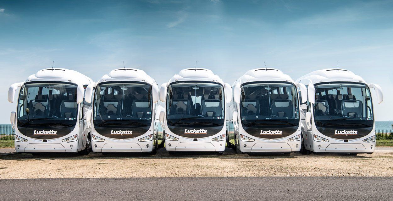 Five white tour buses parked in a row, with the ocean in the background.