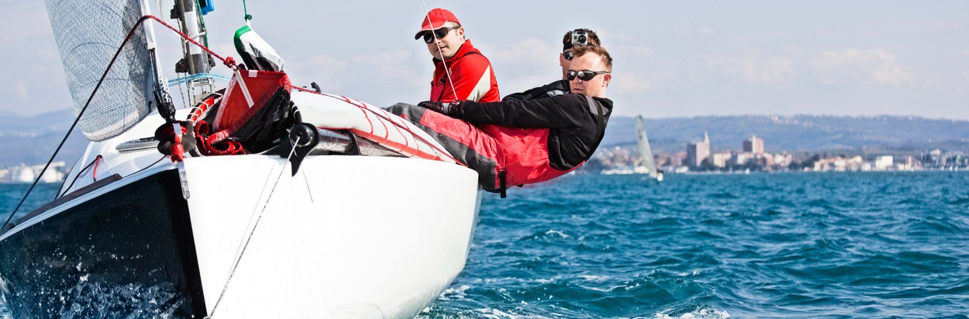 Two people on a sailboat, leaning over the side in the sea. City skyline in the distance.