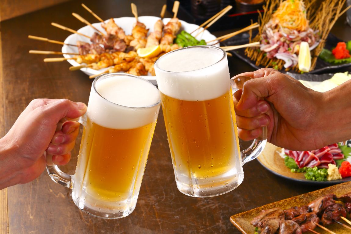Zwei Personen stoßen mit Bierkrügen an, auf einem Tisch stehen japanische Speisen und Getränke.