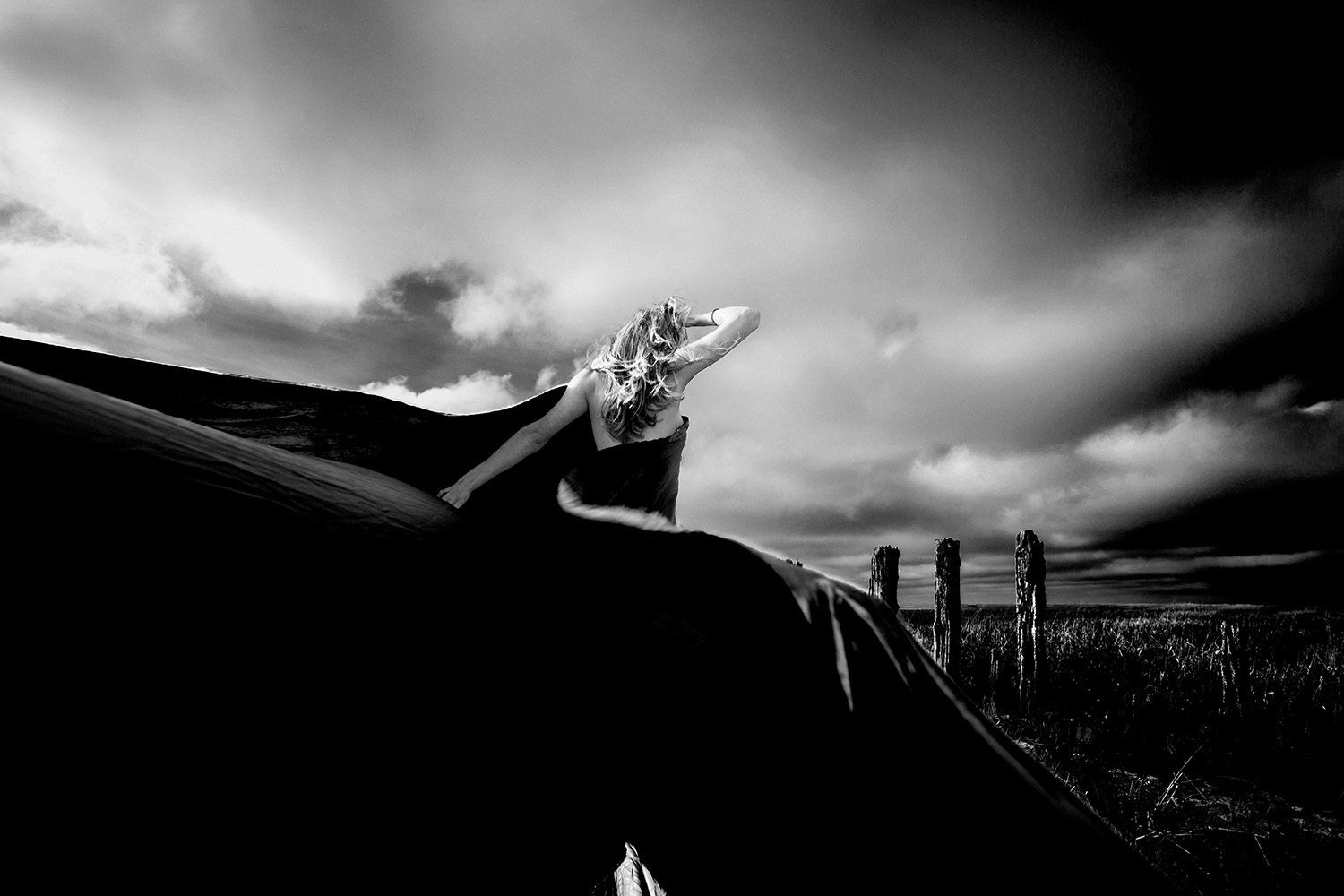 Een vrouw met opgeheven hand zit op een donkere ondergrond en staart naar een dramatische, bewolkte hemel boven een veld.