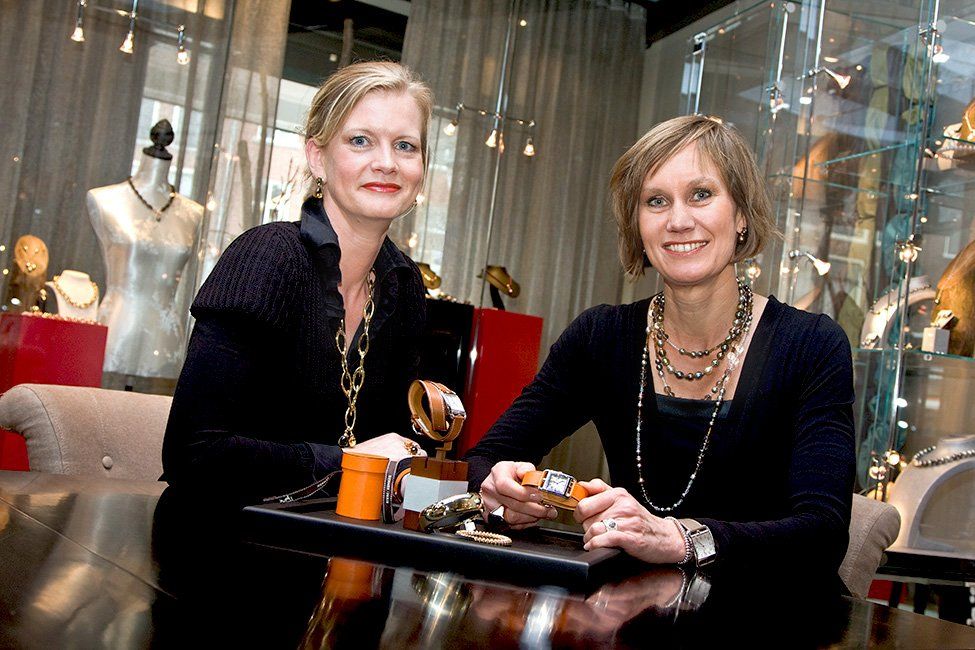 Twee vrouwen staan ​​bij een juweliersbalie en bekijken horloges en halskettingen. Goud, zwart en oranje accenten.