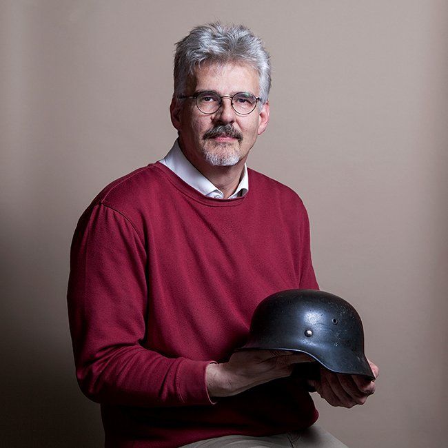 Man in bordeauxrode trui houdt een zwarte helm vast; lichte achtergrond.