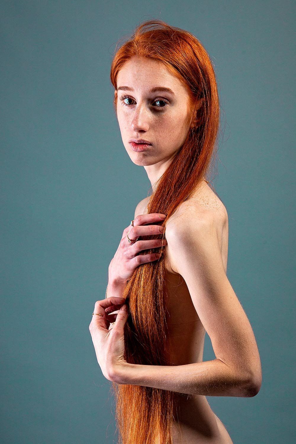 Vrouw met lang rood haar, kijkt in de camera, gedeeltelijk naakt, tegen een turquoise achtergrond.