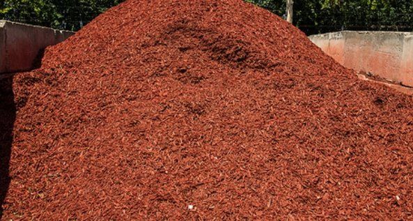 Large pile of reddish-brown wood chips in an outdoor yard or storage area, with concrete walls behind it
