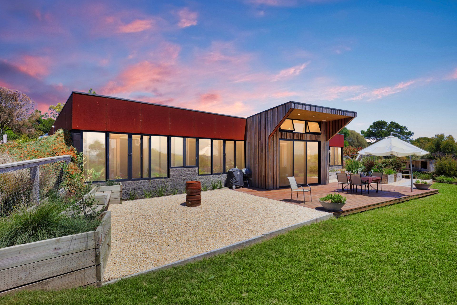 Modern house with red roof, glass walls, and patio at sunset beside a green lawn
